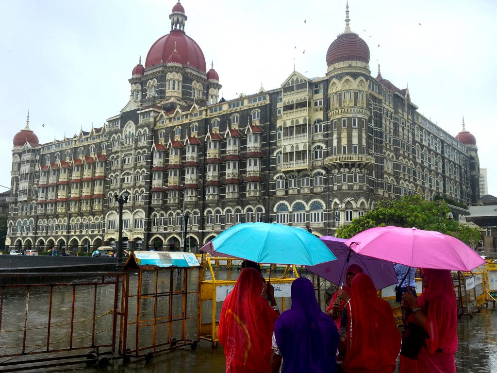 Women in colorful sarees with colorful umbrellas looking at the Taj Mahal Hotel across from the Gateway of India