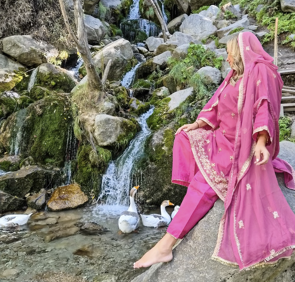 Solo female travel influencer Alyssa Ramos (@mylifesatravelmovie) by a waterfall with geese in Skardu Pakistan